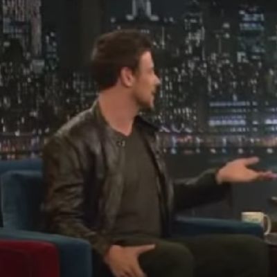 Cory Monteith is wearing a black jacket as he is sitting on a sofa.